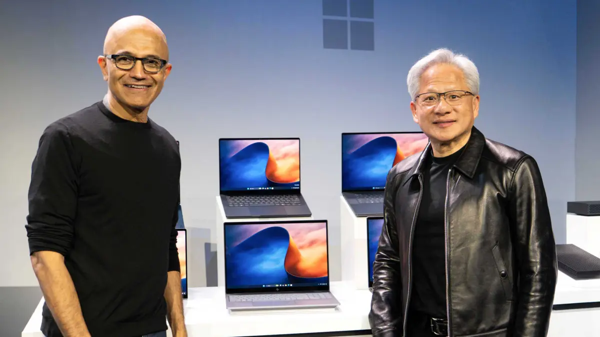 ステージ上で並んで立つ2人のテック企業幹部と、背後の台に展示された複数のノートPC