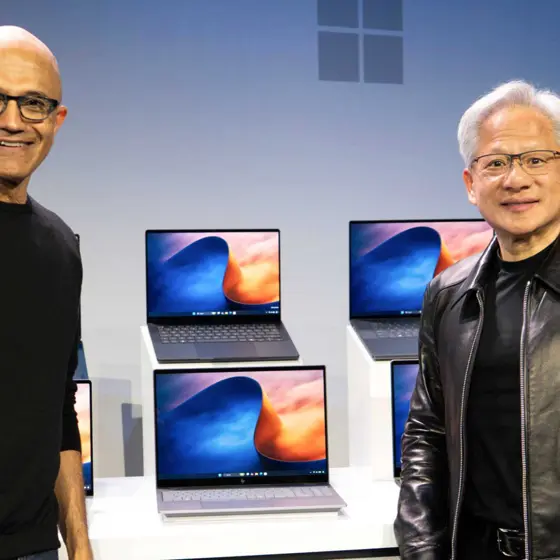ステージ上で並んで立つ2人のテック企業幹部と、背後に展示された複数のノートPC